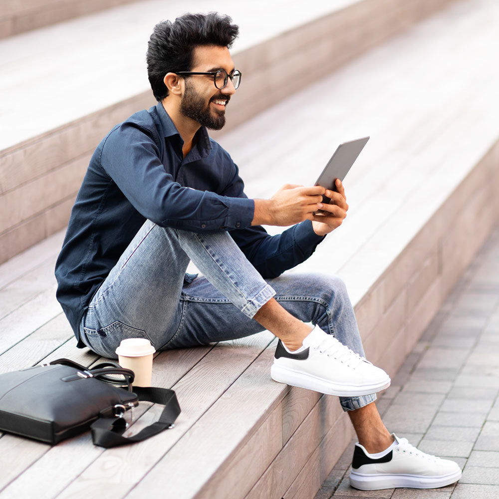 Man sitting on stairs, taking a break from walking. Looking at tablet and drinking coffee while wearing white canvas shoes with Pinnacle 3/4 inside.