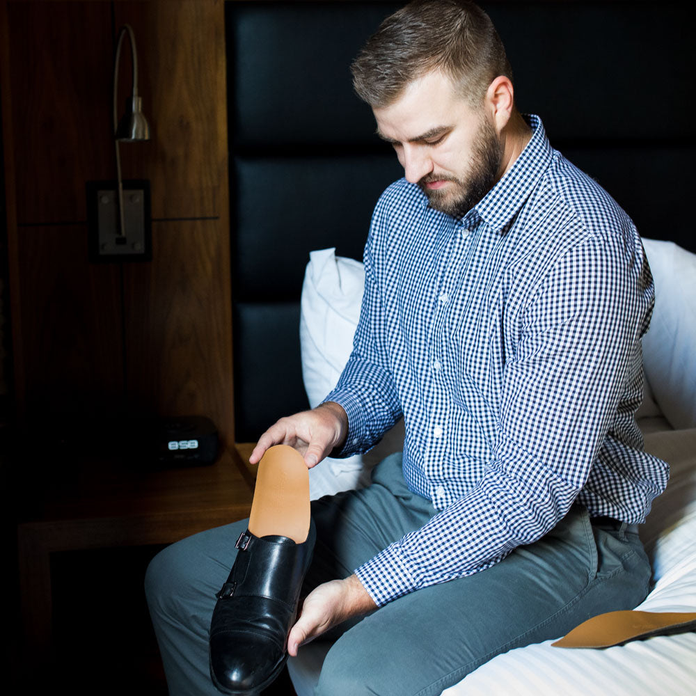 Man sitting on bed getting ready for work, placing PowerStep Pinnacle Dress 3/4 insoles into black leather dress shoes.