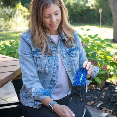 Woman sitting outside at picnic table, placing PowerStep Pinnacle High insoles into black sneakers.