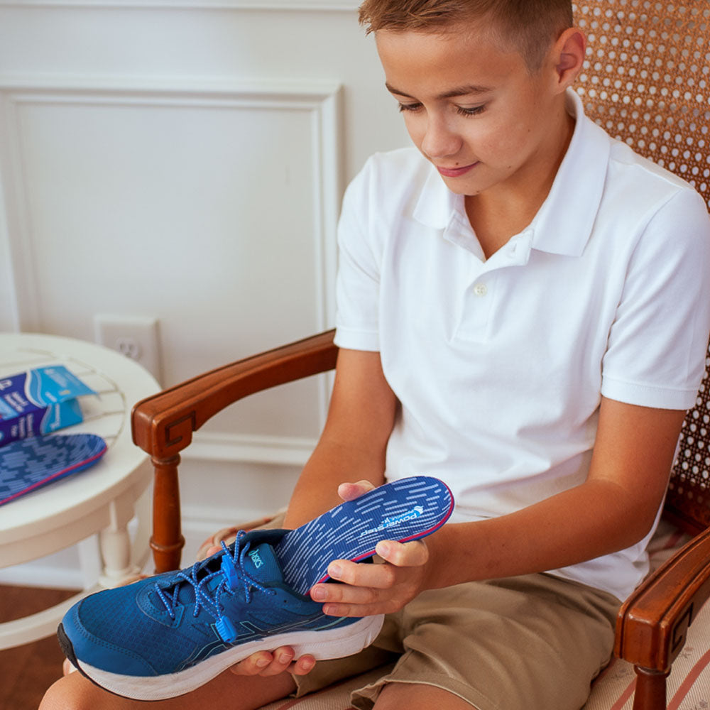 Little boy sitting in chair, placing PowerStep Pinnacle Junior insoles into blue children’s shoe.