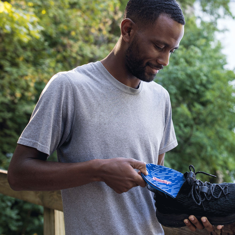 Man standing outside in front of green tree while holding up black tennis shoe and placing PowerStep Pinnacle Plus insoles inside