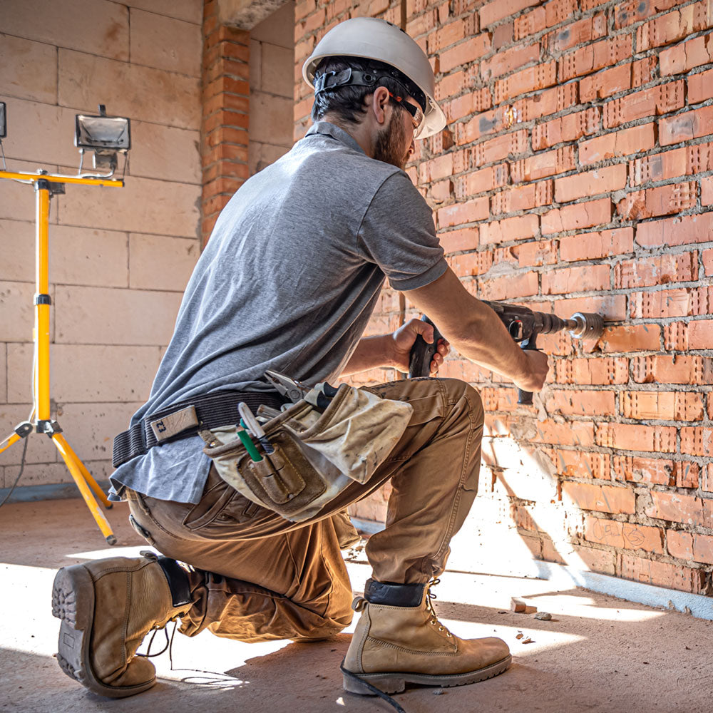 Man in old building wearing hard hat, goggles, and work boots while drilling into brick wall