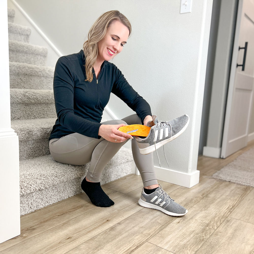 Woman sitting on carpeted stairs while placing PowerStep PULSE Plus insoles into light gray running shoes