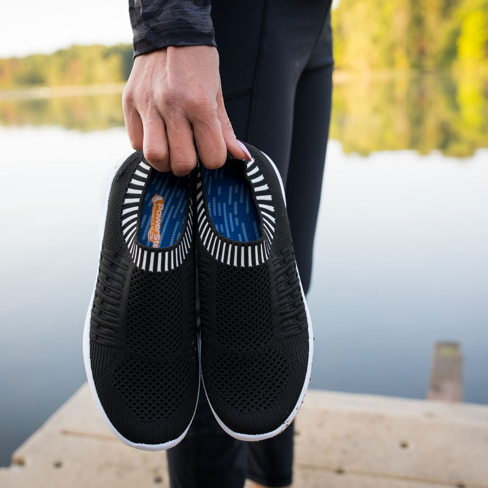 Woman standing on dock in front of lake while holding black and white sneakers with PowerStep Pinnacle Low Insoles inside