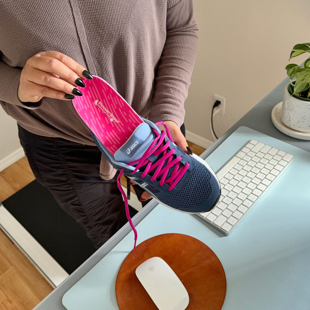 Woman standing at computer desk while holding up blue and pink sneaker to place PowerStep Pinnacle Pink insoles inside