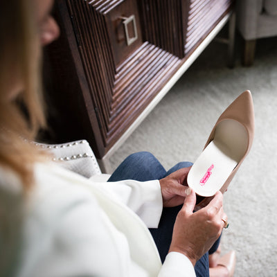 Over-the-shoulder view of woman sitting in bedroom and placing khaki colored PowerStep SlenderFit 3/4 insoles inside of tan high heel shoes #color_khaki