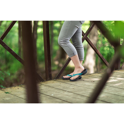 PowerStep Orthotic Arch Supporting Sandals for Women, woman standing on bridge in teal and charcoal sandals with green trees in the background #color_teal-charcoal